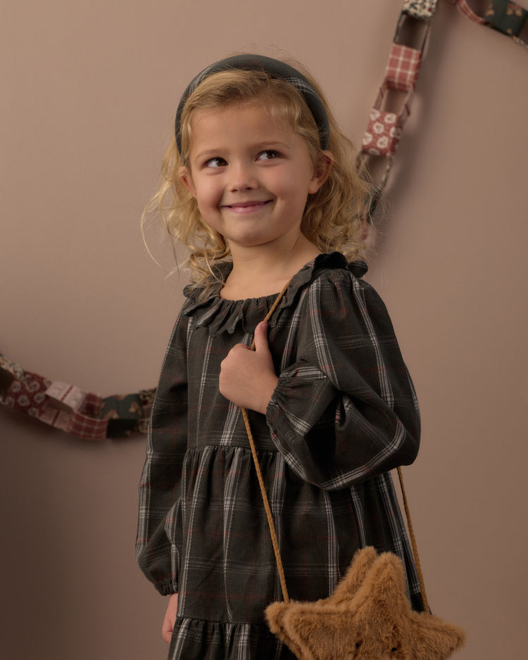 A young girl with blonde curly hair wearing a dark gray plaid long-sleeve dress and a dark headband, holding a brown star-shaped purse against a warm beige background.