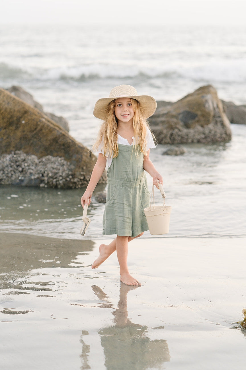 Young girl in a green dress and hat standing on a beach with rocks and water in the background.