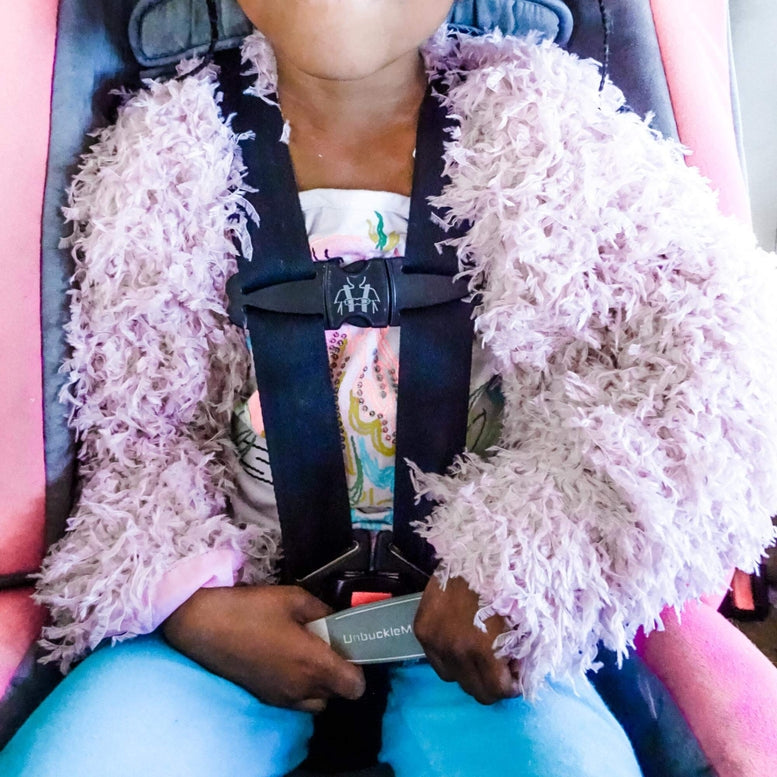 Child in a car seat wearing a fluffy pink jacket, holding an unbuckling device.