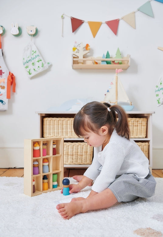 A young child sits on a white rug in a bright, decorated nursery, playing with a wooden toy set containing colorful bottles and small figures.