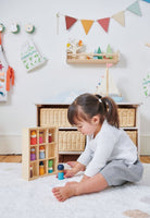 A young child sits on a white rug in a bright, decorated nursery, playing with a wooden toy set containing colorful bottles and small figures.