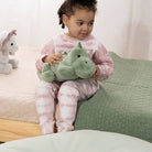 A young child wearing a pink and white tie-dye pajama set sits on a bed, holding a green plush dinosaur toy.
