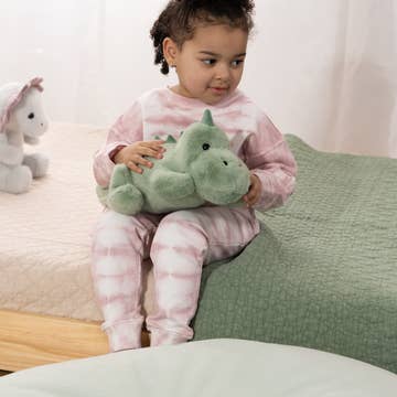 A young child wearing a pink and white tie-dye pajama set sits on a bed, holding a green plush dinosaur toy.