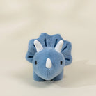 A small blue plush dinosaur toy with white horns and ears, photographed against a plain warm off-white background.