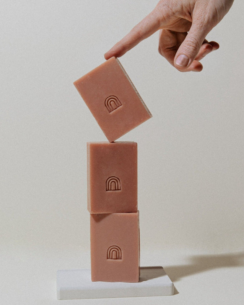 Three dusty pink rectangular bars of soap stacked on a white block, with a hand pointing toward the top bar, against a warm off-white background.