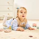 A baby girl lying on a cream carpet in a playroom, wearing a blue and white striped long-sleeve shirt with a tan bib. She is surrounded by pastel-colored wooden building blocks.