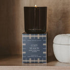 A lit black glass candle sits on top of its box, which reads 'Cozy Season' by Sweet Water Scent. The box has a navy and beige plaid pattern and is displayed on a wooden shelf next to a white decorative pumpkin against a warm brown background.