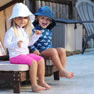 Two children sitting on a wooden bench wearing sun hats and colorful clothing.
