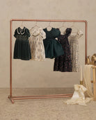 A copper-colored clothing rack displaying five vintage-style children's dresses in various colors and floral patterns, set against a neutral textured beige wall and floor.