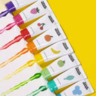 Eight white paint tubes with colorful fruit illustrations and black '$$$$$' text, arranged diagonally on a bright yellow background with corresponding paint splashes visible.