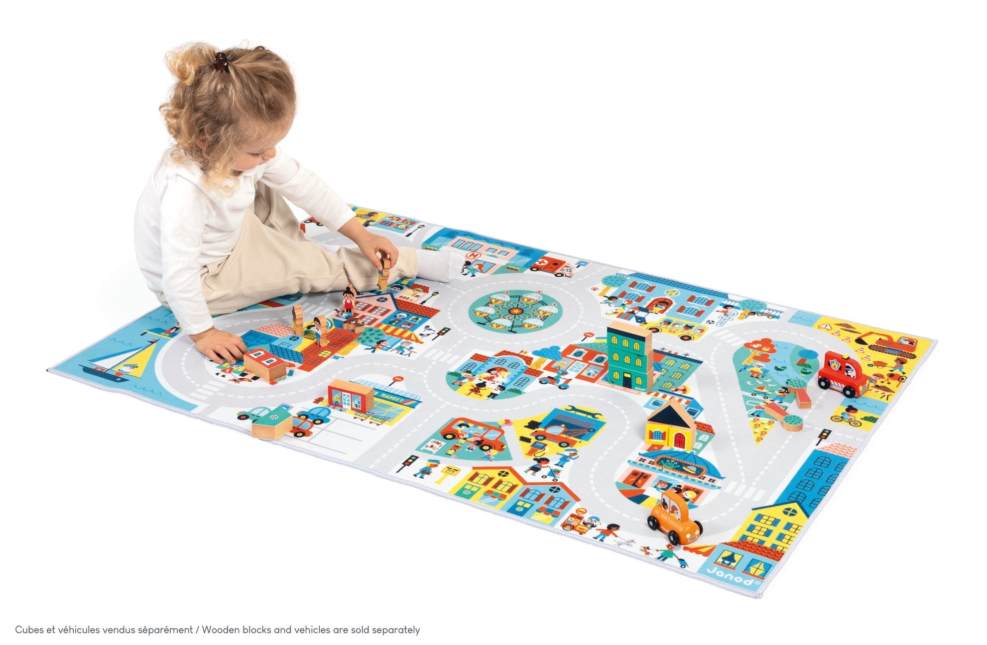 A young child sits on a colorful city play mat with toy cars and blocks on a clean white background. The play mat features illustrated roads, buildings, and various play pieces.
