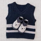 A baby knit sweater vest in navy blue with two horizontal white stripes and a white bootie with a navy cuff and embroidered bear face, laid flat on a textured beige fabric background.