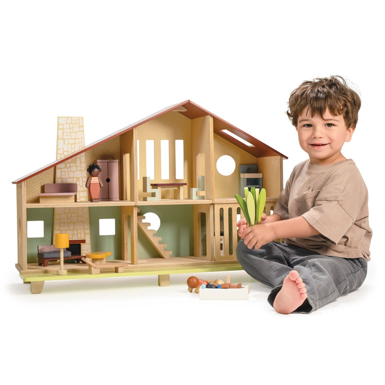 A smiling young child sits cross-legged on the floor playing with the Wooden Cedar Chalet Modernist Dollhouse by Mentari, holding toy vegetables while dolls and furnishings are arranged inside and outside the open-front mid-century modern home.