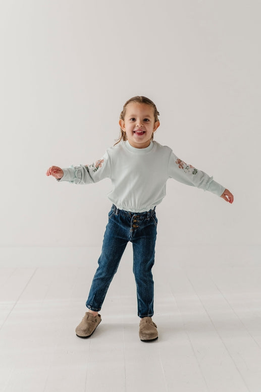 A young child wearing a light sage green long-sleeve crewneck sweatshirt with floral embroidery on the sleeves, paired with blue jeans and brown suede boots, standing with arms outstretched against a white studio background.