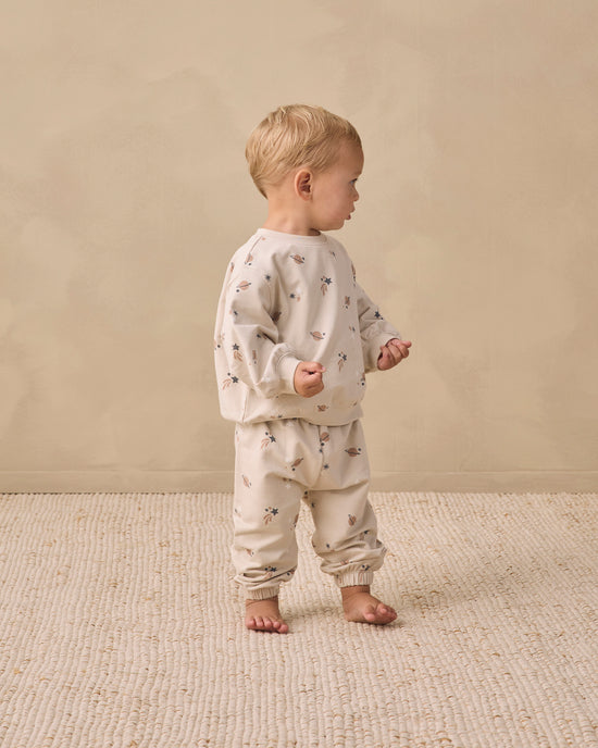 A toddler stands barefoot in profile wearing a light beige two-piece pajama set with a small scattered floral print, photographed against a warm beige wall and textured rug.