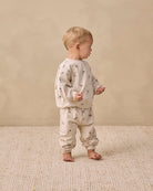 A toddler stands barefoot in profile wearing a light beige two-piece pajama set with a small scattered floral print, photographed against a warm beige wall and textured rug.