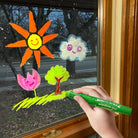 Hand holding a green window gel crayon with colorful window decorations on a glass door.