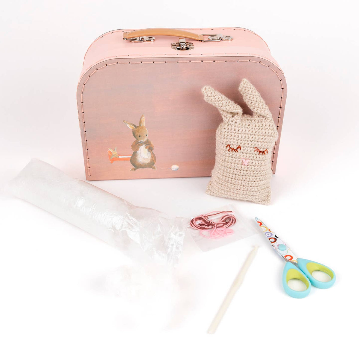 Pink suitcase with a crocheted bunny, scissors, and other crafting items on a white background