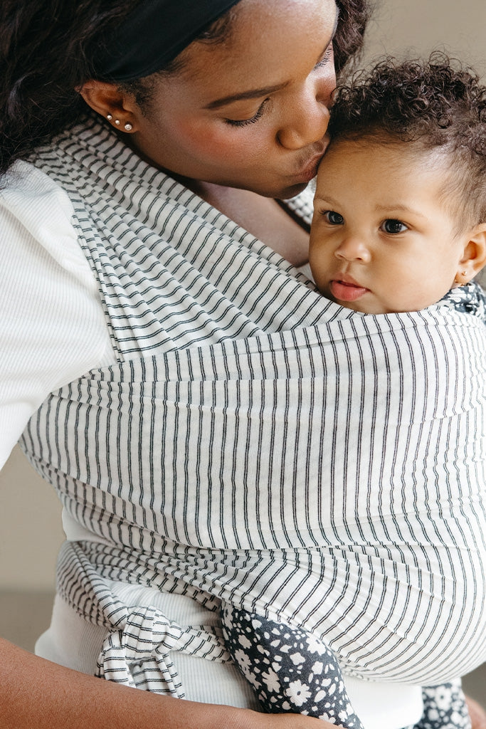 Woman holding a baby in a striped baby carrier against a neutral background