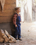 A young boy wearing blue denim overalls over a brown and orange striped long-sleeve shirt stands outdoors next to a rustic wooden wall with stacked firewood.
