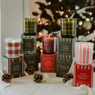 A collection of Christmas-themed scented candles in glass jars with plaid and striped patterns, displayed on top of their branded gift boxes on a white textured blanket with pinecones and a blurred Christmas tree in the background.