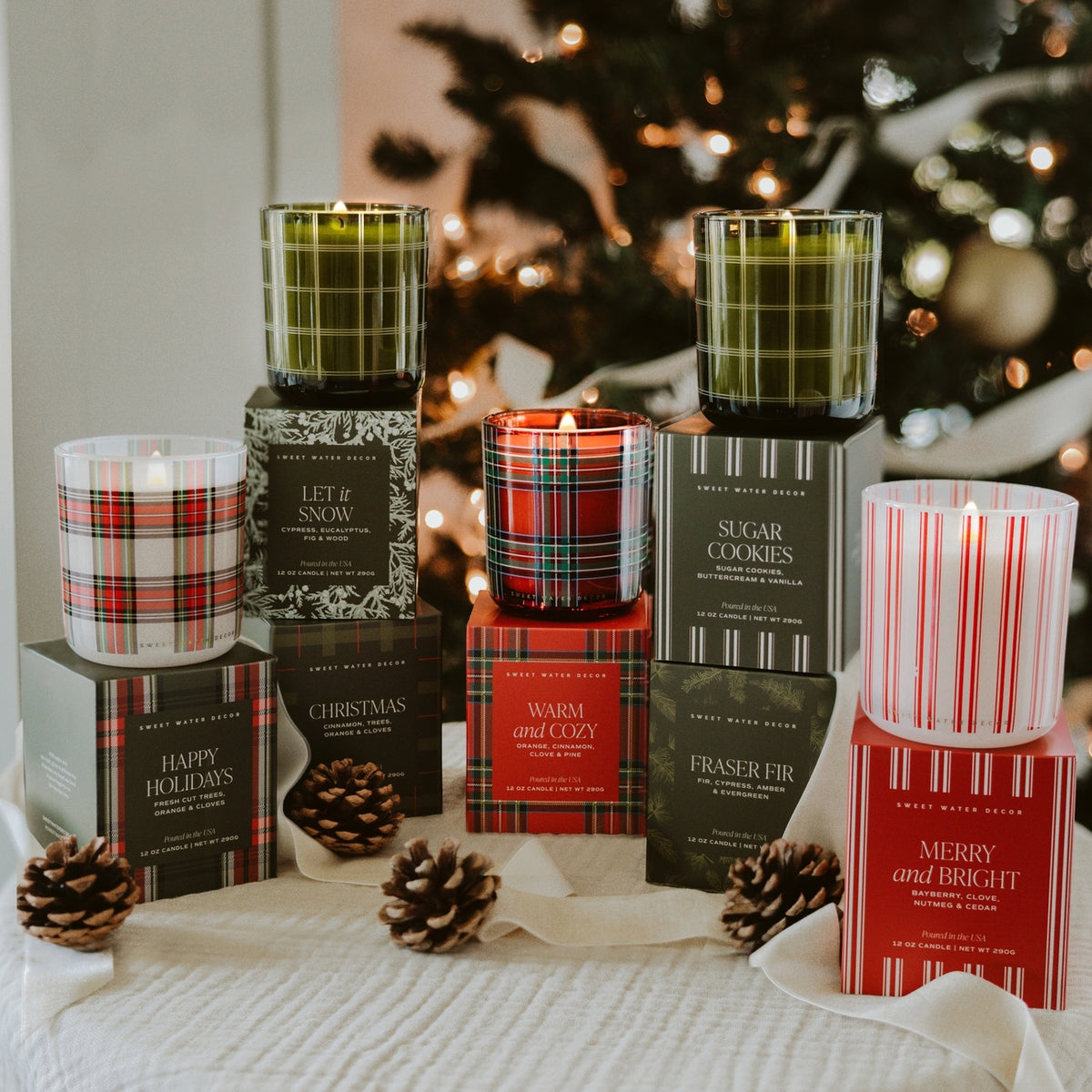 A collection of Christmas-themed scented candles in glass jars with plaid and striped patterns, displayed on top of their branded gift boxes on a white textured blanket with pinecones and a blurred Christmas tree in the background.