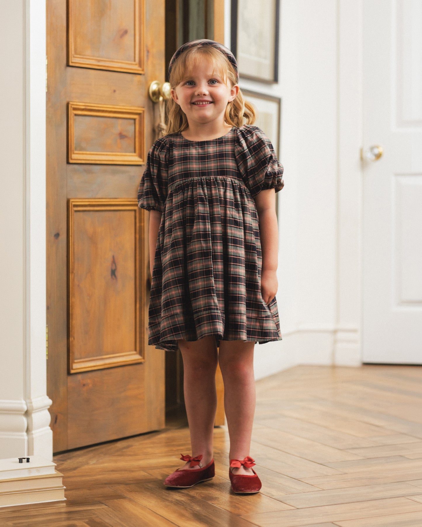 A young girl stands in a doorway wearing a short-sleeve plaid dress with puff sleeves, paired with red shoes and a headband. The dress has a dark brown and black plaid pattern.