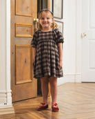A young girl stands in a doorway wearing a short-sleeve plaid dress with puff sleeves, paired with red shoes and a headband. The dress has a dark brown and black plaid pattern.