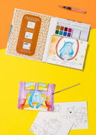 A children's watercolor painting kit shown open on a bright yellow and orange background, with coloring pages, a paintbrush, and a watercolor palette visible. The kit includes printed coloring pages and a painted illustration of a blue monster.