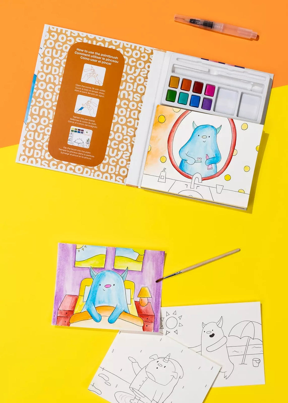 A children's watercolor painting kit shown open on a bright yellow and orange background, with coloring pages, a paintbrush, and a watercolor palette visible. The kit includes printed coloring pages and a painted illustration of a blue monster.