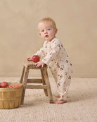 A baby standing on a woven rug and leaning on a rustic wooden stool, wearing a cream footed sleeper with a brown cherry print. A small wooden basket with red apples sits on the floor to the left.