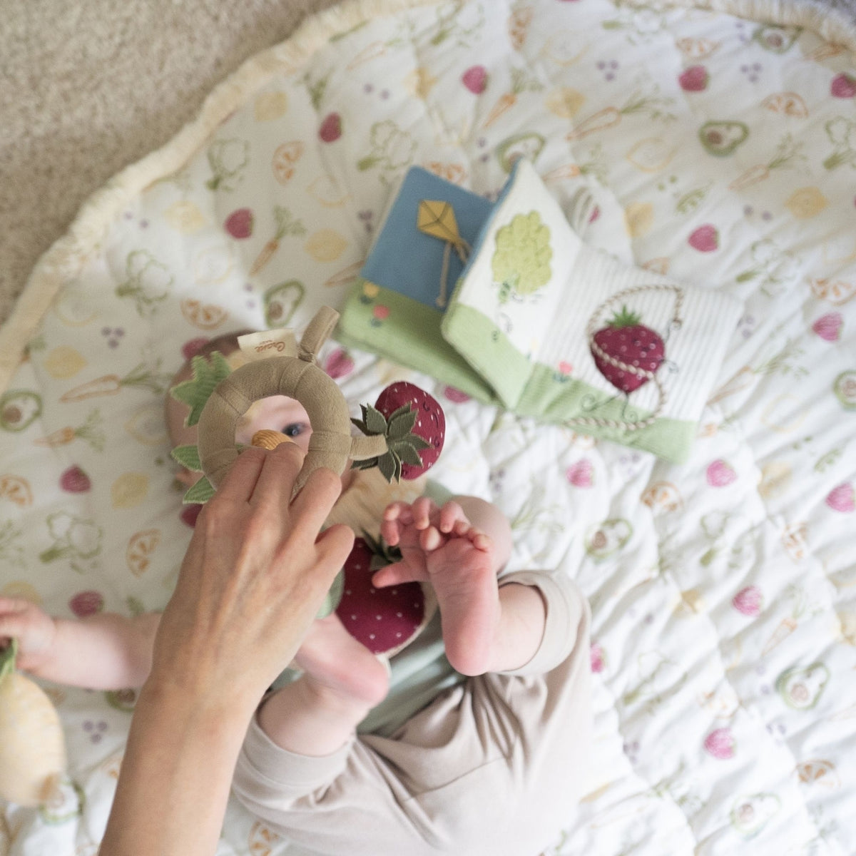 A baby lying on a round quilted play mat with a strawberry and fruit pattern, interacting with a plush animal toy. A matching book and pillow are placed nearby on the mat.