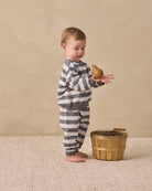 A toddler stands barefoot on a textured cream rug, wearing a gray and white striped long-sleeve pajama set and holding a small brown pear. A small wooden bucket sits beside them against a warm beige studio backdrop.