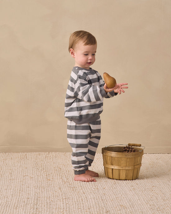 A toddler stands barefoot on a textured cream rug, wearing a gray and white striped long-sleeve pajama set and holding a small brown pear. A small wooden bucket sits beside them against a warm beige studio backdrop.