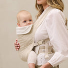Woman holding a baby in a beige baby carrier against a plain background