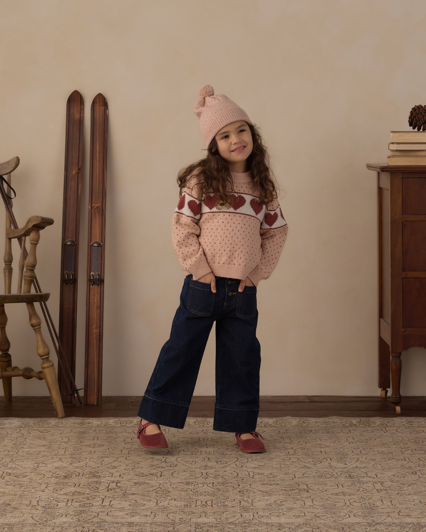 A young girl stands in a cozy indoor room wearing a pink knit beanie, a pink patterned sweater, dark blue wide-leg jeans, and pink shoes. The background features a beige wall, vintage wooden skis, a wooden chair, and a wooden cabinet.