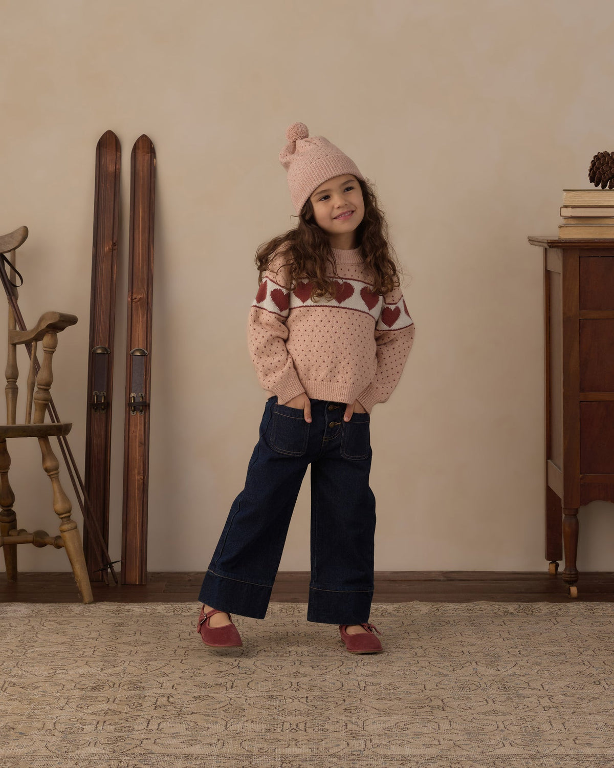A young girl stands in a cozy indoor room wearing a pink knit beanie, a pink patterned sweater, dark blue wide-leg jeans, and pink shoes. The background features a beige wall, vintage wooden skis, a wooden chair, and a wooden cabinet.