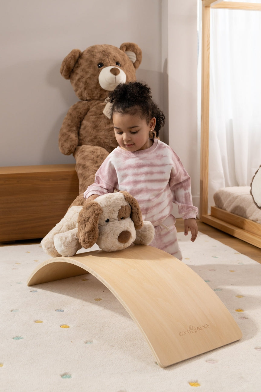 A toddler in pink striped pajamas stands on a curved wooden toy ramp in a child's bedroom, holding a plush dog toy. A large teddy bear sits in the background near a bed.