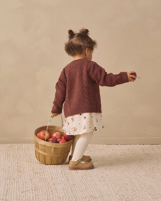 A toddler stands with their back to the camera, wearing a burgundy cable-knit cardigan over a white floral dress, white tights, and tan shoes. They are holding a small wooden basket filled with red apples against a warm beige background.