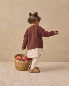 A toddler stands with their back to the camera, wearing a burgundy cable-knit cardigan over a white floral dress, white tights, and tan shoes. They are holding a small wooden basket filled with red apples against a warm beige background.