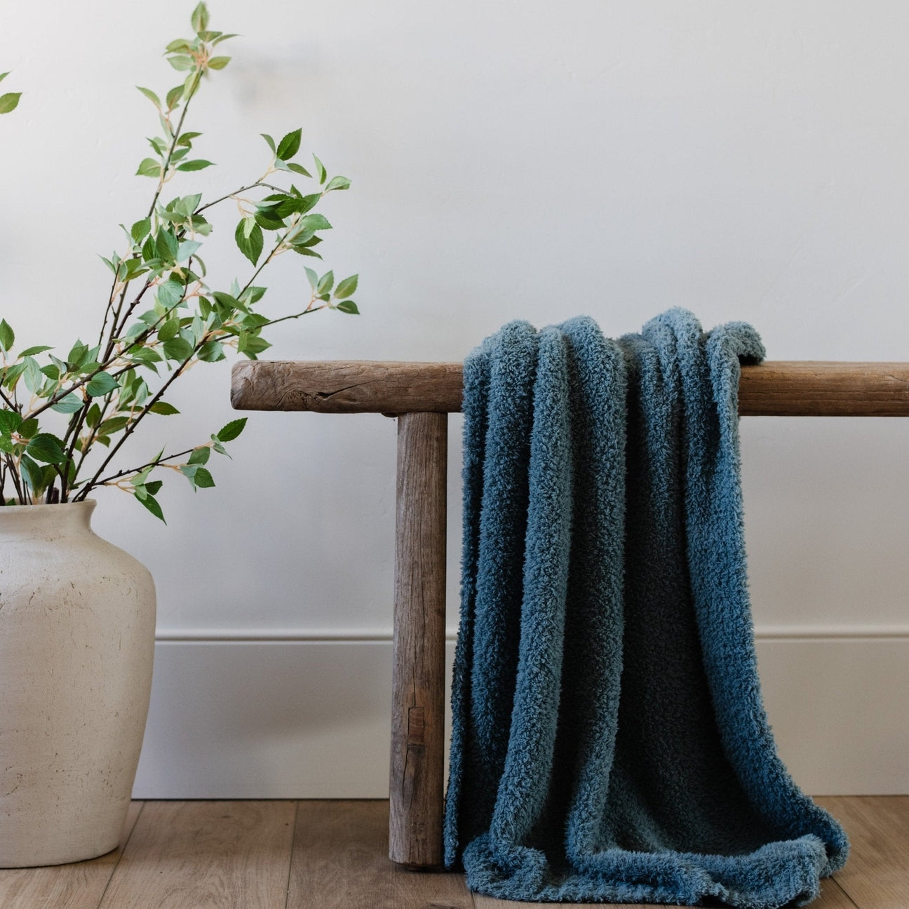 A soft, teal-colored textured blanket draped over a wooden bench next to a vase with greenery