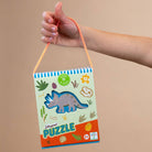 Hand holding a dinosaur-themed shaped puzzle packaging against a beige background
