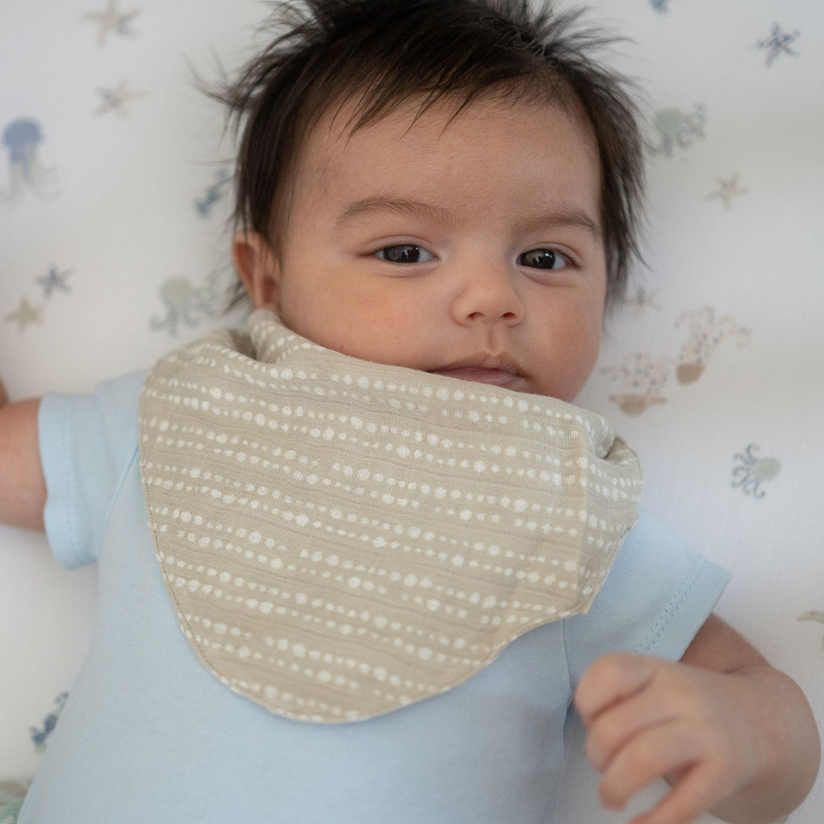 Baby wearing a light blue onesie with a beige bib, lying on a white blanket with blue patterns.