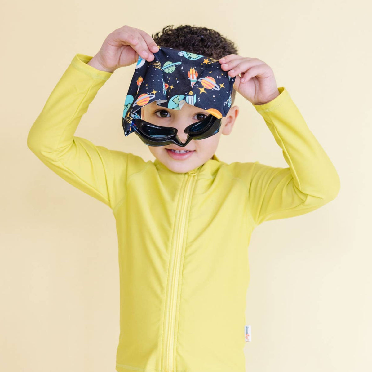 Child wearing a yellow jacket and colorful space-themed goggles against a beige background