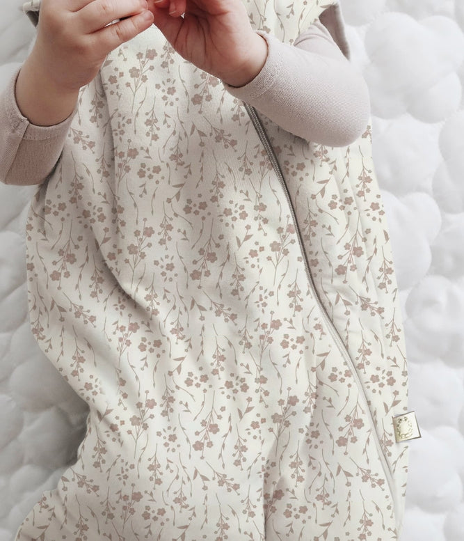 A close-up of a baby wearing a cream-colored quilted sleep sack with a delicate brown floral pattern, layered over a light beige long-sleeve shirt, lying on a white quilted mattress.