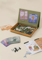 An educational animal-themed board game set displayed on a light beige background, featuring an olive green box with an illustrated interior, animal cards, wooden animal pieces, and an instruction booklet.