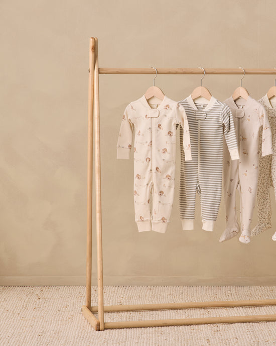 Four baby footed sleepers in neutral tones hanging on a wooden clothing rack against a textured beige wall and floor.