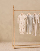 Four baby footed sleepers in neutral tones hanging on a wooden clothing rack against a textured beige wall and floor.