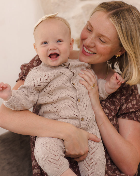 A baby wearing a beige knit button-front romper and matching headband is held by a smiling woman in a brown floral dress in a warm indoor setting.