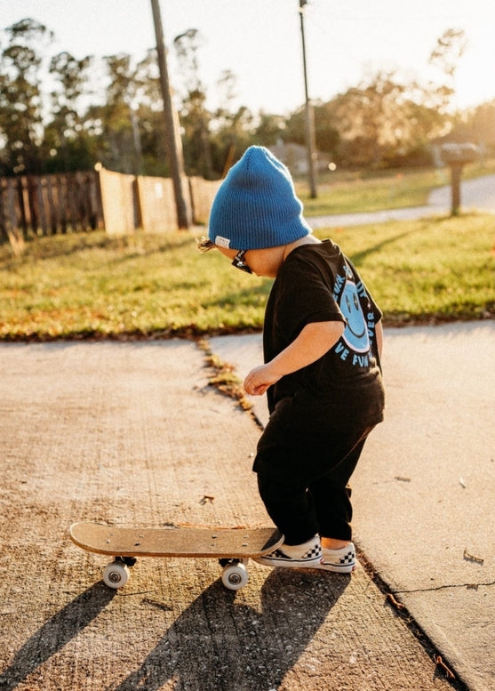 Child wearing a blue beanie and black shirt with text, standing on a skateboard outdoors.